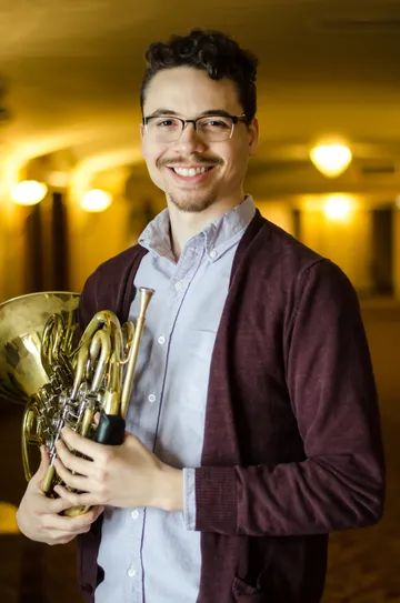 Tyler, low-20s with mustache and short beard, looking directly into the camera, holding a french horn with a blue button up shirt and red cardigan standing in a blurry hallway at Eastman School of Music.