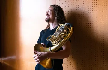 Tyler with long dark hair and goatee, wearing a black vneck shirt and holding a french horn leaning against a peg board wall and looking off to the left, smiling.