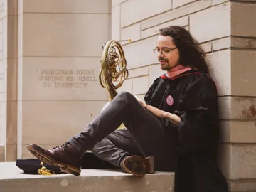 Tyler, in a graduation robes, with long dark hair, sits on a stone wall with his back against the outside of a university building holding a french horn by the bell.