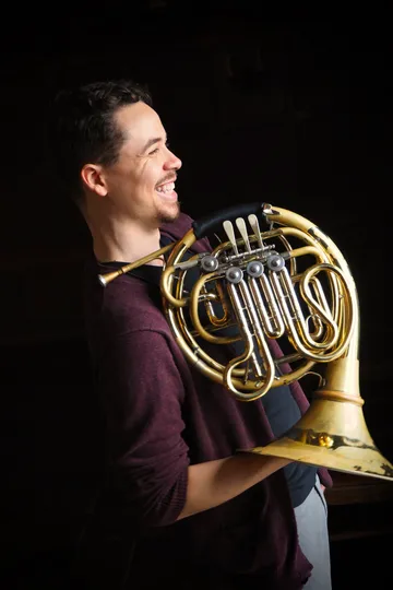 Tyler, in profile looking to the right, in his mid-20s, wearing a black v-neck shirt and a maroon cardigan, holding a french horn with his hand in the bell, smiling ebulliently.